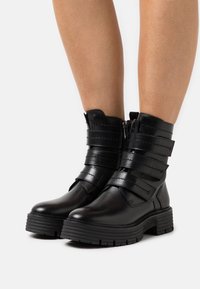 Bottines noires en cuir avec plusieurs sangles horizontales, un bout arrondi, une semelle épaisse et une fermeture éclair sur le côté pour un enfilage facile.