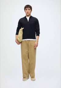 Ung mann i svart genser med halvlange glidelås, beige vidbukser og beige sko, holder en beige sammenbrettet jakke mot hvit bakgrunn.