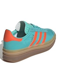 Zelene in oranžne superge Adidas Gazelle z gladkim zgornjim delom in teksturirano peto. Ponuja tri oranžne stripe in prosojen podplat.