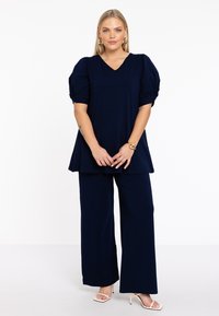 Navy-blauwe top met v-hals en pofmouwen, gecombineerd met bijpassende wijde broek. Gemaakt van een gladde, zachte stof. Sleehakken maken de look compleet.