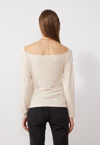 Ljus beige, off-the-shoulder topp med långa ärmar. Mjuk textur och figurnära design, med en subtil detalj på baksidan med en kedja. Svarta byxor.