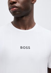Κοντινό πλάνο ενός ατόμου που φοράει ένα λευκό T-shirt με τη λέξη «BOSS» τυπωμένη γκρι στο στήθος.