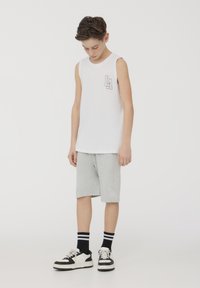 Camicetta bianca senza maniche in cotone con logo viola sul petto, abbinata a pantaloni corti grigi con logo, scarpe nere e bianche, e calzini neri.