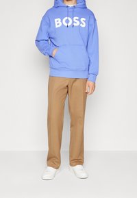 Ljusblå huvtröja med "BOSS" i vita bokstäver, framficka, kombinerad med beige byxor och vita sneakers. Enkel, avslappnad design.
