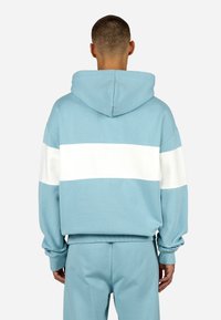 Modrá mikina s kapucňou a bielym horizontálnym pruhom na chrbte, s voľným strihom a rebrovanými manžetami a spodným okrajom.