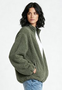 Donna con capelli neri e ondulati che indossa una giacca sherpa verde oliva oversize, camicia bianca e jeans blu, in piedi con le mani nelle tasche.