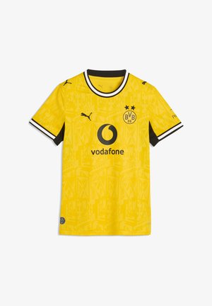 Žena v žltom drese Borussie Dortmund drží čierno-žltú futbalovú loptu, stojí pred sivým pozadím.