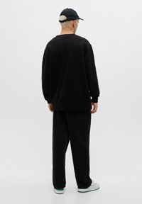 Svart oversized sweatshirt och matchande byxor i mjukt tyg. Tillbehören inkluderar en svart keps och vita sneakers med gröna accenter.