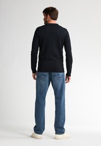 Tricou negru cu mânecă lungă, cu textură cordonată orizontal, asociat cu blugi decolorati de un albastru deschis și adidași albi. Vedere din spate.