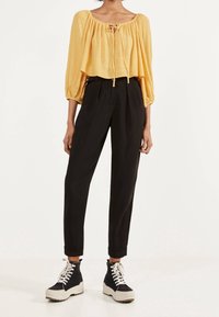 Żółta skrócona bluzka z marszczonymi rękawami i wiązaniem z przodu; zestawiona z czarnymi spodniami w wysokim stanie z fałdami i czarnymi botkami za kostkę.