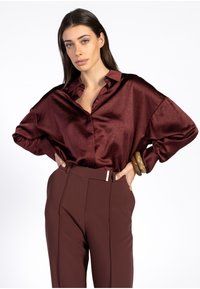 Vrouw met lang donker haar draagt een losse, glanzende bordeauxrode blouse en hoge bruine broeken, poseert met handen op de heupen tegen een witte achtergrond.