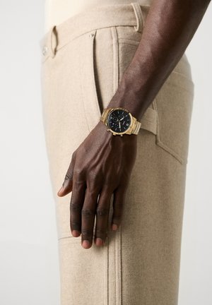 Main à la peau foncée reposant près de la poche d'un pantalon beige, portant une montre-bracelet en or avec un cadran noir et un bracelet métallique.
