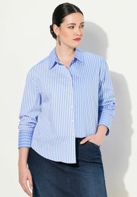 Donna con capelli scuri, indossante una camicia a righe verticali blu e bianche e una gonna di jeans scura, che guarda di lato con una mano in tasca.
