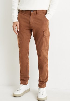 Mężczyzna w rdzawej odzieży typu cargo z bocznymi kieszeniami, białych trampkach i białej bluzie z długim rękawem, stojący na jednolitym tle.