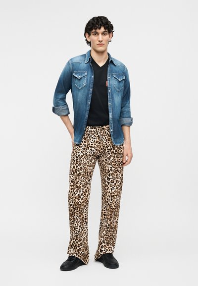 Άνδρας με πουκάμισο τζιν με ανεβασμένα μανίκια, μαύρο μπλουζάκι λαιμόκοψης σε σχήμα V, παντελόνι με λεοπάρ σχέδιο και μαύρα παπούτσια, στέκεται μπροστά σε απλό φόντο.