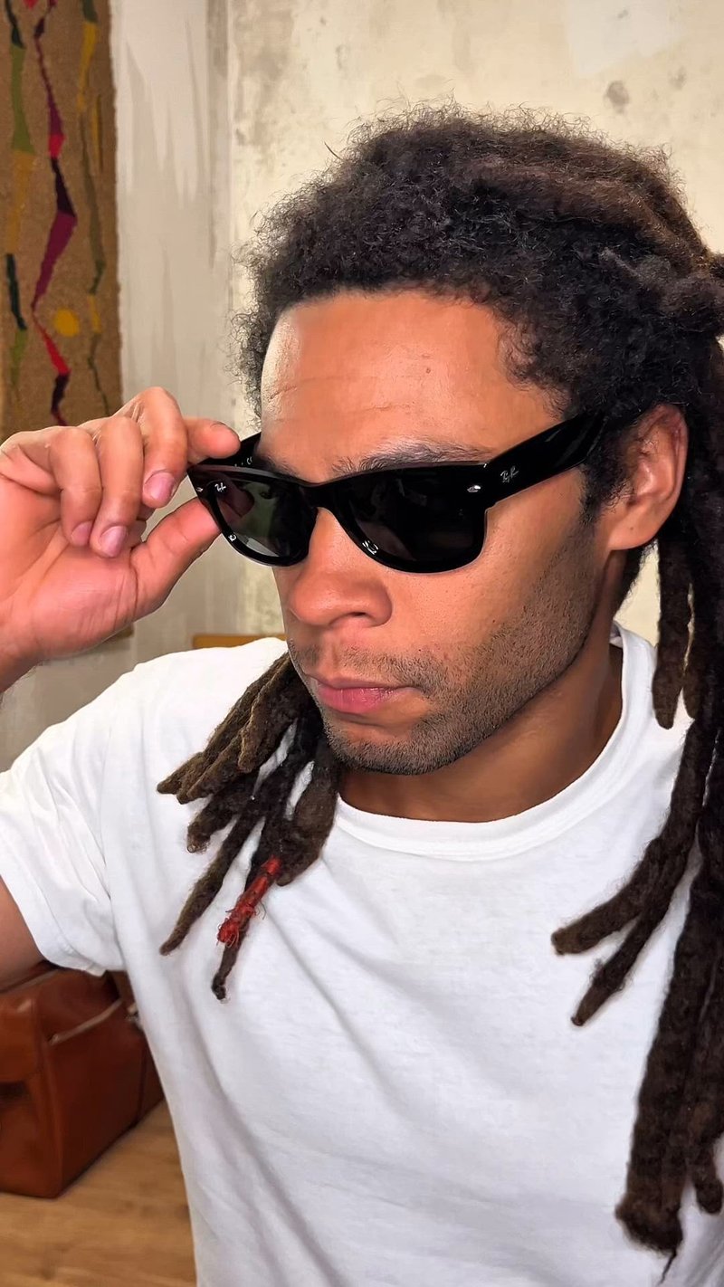 Мъж с дълги дредове, който коригира черни слънчеви очила Ray-Ban, облечен в бяла тениска, с текстурирана стена и шарени завеси на заден план.