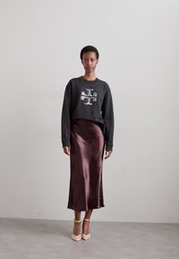 Μαύρη ζακέτα με λογότυπο από παγιέτες, συνδυασμένη με μια ρέουσα σκούρα καφέ σατέν φούστα. Το μοντέλο φοράει γυμνά πέδιλα με λουράκια. Απλό, καθαρό φόντο.