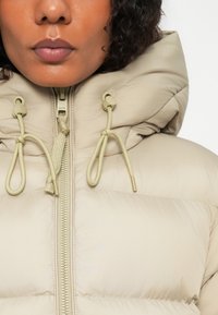 Chaqueta acolchada beige con cuello alto, que presenta un cierre delantero con cremallera y cordón ajustable. Textura suave, diseño ligero y acentos mínimos.