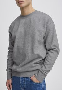 Mand iført en enkel grå langærmet crewneck-sweatshirt med hænderne i lommerne på blå jeans mod en ensartet lys baggrund.