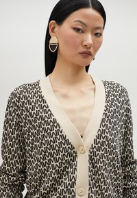 Pleteni kardigan u bež i smeškim bojama, s geometrijskim uzorkom, V-izrezom i dva prednja dugmeta. Model nosi velike okrugle naušnice.