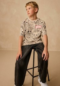 Beige t-shirt med prints af Marvel-tegnefilmfigurer, korte ærmer og rødt logo. Sorte løse jeans og hvide sneakers fuldender outfittet.