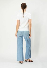 Valkoinen lyhythihainen paita, jossa on koristeelliset takanapit, yhdistettynä vaaleansinisiin leveälahkeisiin farkkuihin ja mustiin litteisiin sandaaleihin.