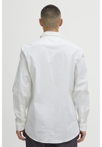 Camisa de algodón blanca con mangas largas, puños abotonados, dobladillo curvado y cuello clásico. Textura suave sin patrones visibles.