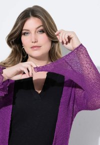 Lila öppningsstickad cardigan med långa ärmar, lagd över en svart V-ringad topp. Cardiganen har en texturerad, lätt design.