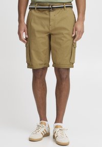 Shorts cargo kaki avec poches latérales, ourlets roulés et une ceinture rayée contrastante. Portés avec des baskets blanches et beiges.