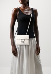 Hvid skuldertaske af læder med sølvnitter, rektangulær form, klaplås og justerbar rem. Kombineret med en sort tanktop og hvid nederdel.