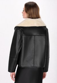 Schwarze Lederjacke mit einem krämfarbenen Schurwollkragen. Die Jacke hat eine kastige Form, gerade geschnittene Ärmel und eine glatte Textur.