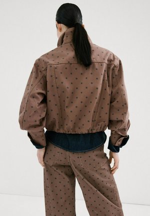 Personne avec une queue de cheval noire portant une veste marron à pois et un pantalon assorti, chemise foncée visible à la taille et aux poignets, debout de dos.