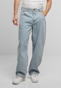 Helle blaue, weit geschnittene Jeans aus Denim, ausgestattet mit fünf Taschen, einem Reißverschluss und einem gelben Etikett an der Gesäßtasche. Kombiniert mit weißen Turnschuhen.