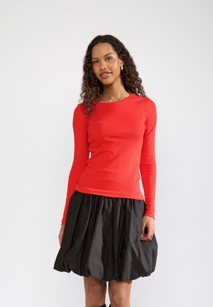 Mujer con cabello rizado que lleva una camiseta roja ajustada de manga larga y una falda negra abullonada, de pie frente a un fondo claro y liso.