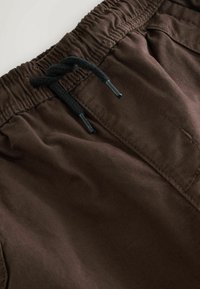 Close-up of brown fabric pants with elastic waistband and black drawstring tied in a knot.