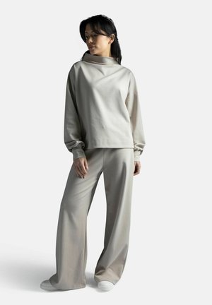 Nainen, joka pukeutunut väljään beigeen collegepuseroon ja leveälahkeisiin housuihin sekä valkoisiin tennareihin, seisoo yksivärisen taustan edessä.