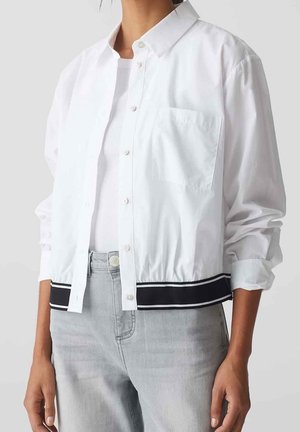 Personne portant une veste blanche boutonnée avec une poche poitrine et un ourlet rayé noir, associée à un jean gris clair et un haut blanc.
