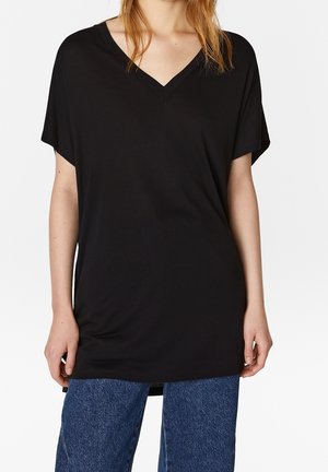 Kvinde iført en løs sort V-hals kortærmet skjorte og blå jeans, stående mod en ensfarvet hvid baggrund.