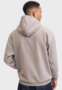 Mann in einem hellgrauen Fleece-Hoodie, von hinten gesehen, vor einem schlichten hellen Hintergrund stehend.