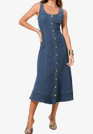 Frau trägt ärmelloses blaues Denim-Midikleid mit durchgeknöpftem Vorderteil und offenen High Heels mit Schlangenhautmuster, posiert mit einer Hand in der Hüfte.