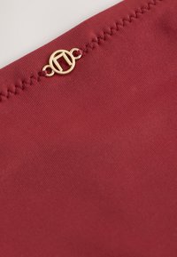 Gros plan sur un tissu bordeaux avec une couture en zigzag et un petit emblème métallique doré rond attaché près du bord.