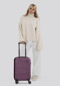 Lila Hartschalentrolley mit vertikalen Rillen, ausziehbarem Griff und Rollen, platziert neben einer Person in einem cremefarbenen Sweater.