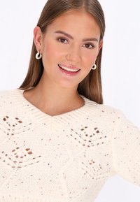 Cremfarbener Strickpullover mit V-Ausschnitt, der mit floralen Mustern und mehrfarbigen Sprenkeln versehen ist. Dazu runde Ohrringe.