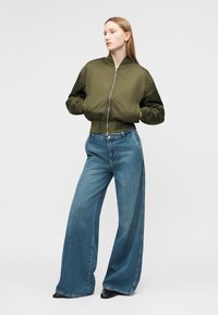 Nő olívazöld, rövidített bomber dzsekiben, bő szárú kék farmerral és fekete cipőben, a kezét a dzseki zsebében tartva áll.