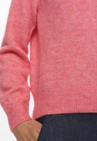 Rozā adīta džemperis ar mīkstu tekstūru, ar ribotām piedurknēm un apakšmalu, kas izceļas ar brīvu piegriezumu. Fona attēls rāda tumšus džinsus.