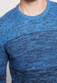 Pull en tricot bleu avec un motif texturé et un col rond. Le tissu présente un mélange de teintes de bleu claires et foncées.