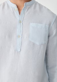 Uomo che indossa una camicia di lino azzurro chiaro con colletto alla coreana, tre bottoni e taschino sul petto, mostrato dal collo alla vita.