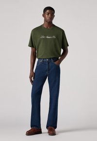 Joven vestido con una camiseta oliva de Levi Strauss & Co., jeans azules y zapatos de cuero marrón, de pie con una mano en el bolsillo.