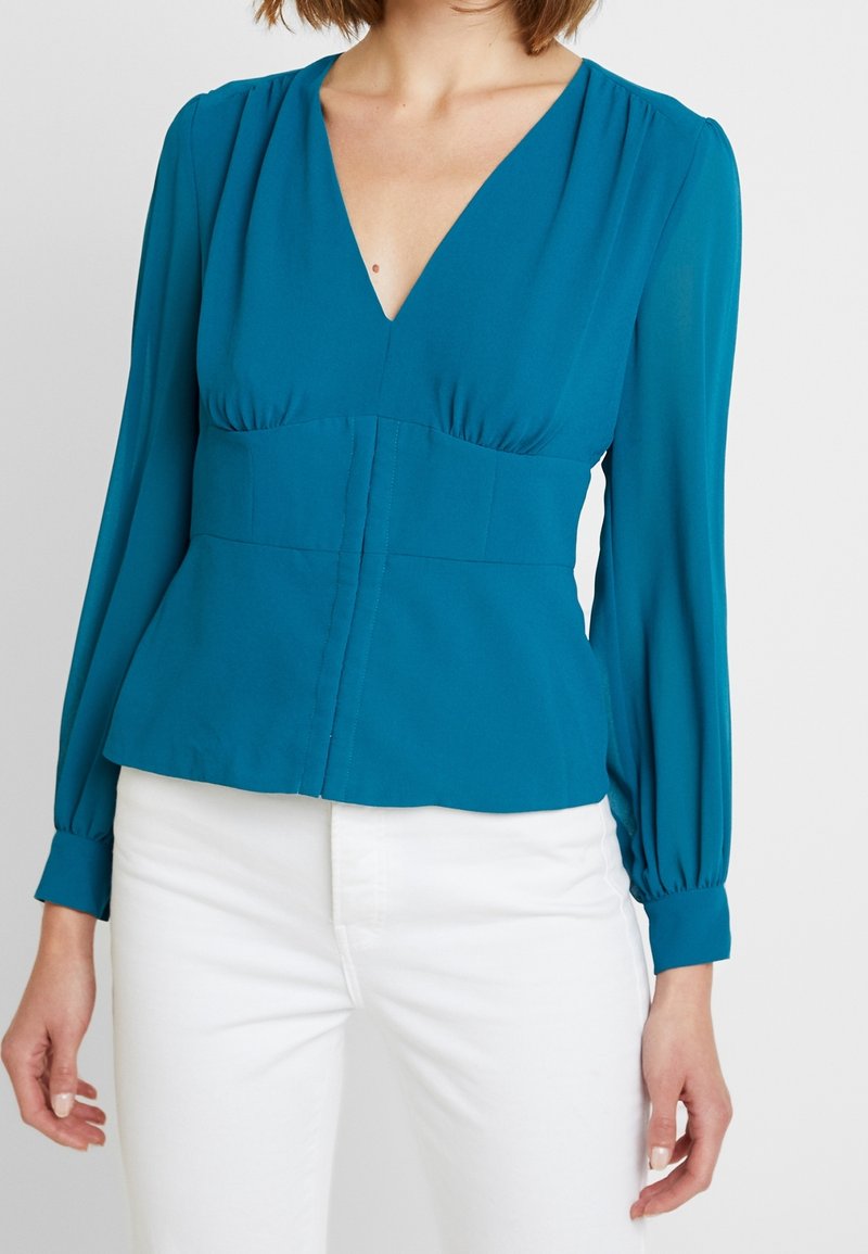Blouse teal avec un décolleté en V profond, des manches bouffantes longues, un corps froncé et une taille ajustée qui s'évase, fabriquée en tissu léger.