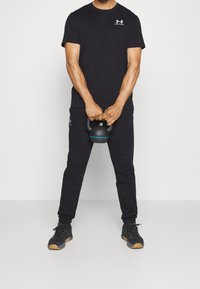 Maglietta atletica nera con logo Under Armour, abbinata a pantaloni della tuta neri. Persona tiene un kettlebell con dettagli blu. Scarpe da ginnastica nere.